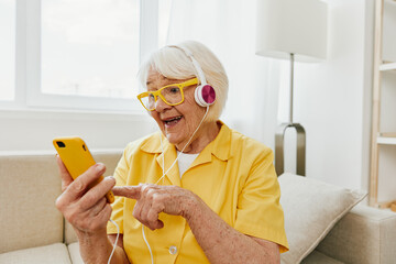 Happy elderly woman watching video call on phone and listening to music on headphones, surprise and open mouth, bright modern interior, lifestyle online communication.