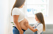 © Louis-Paul Photo - Excited little daughter touching pregnant mom tummy, standing next to window