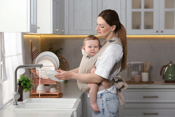 Wall Mural - Mother holding her child in sling (baby carrier) while washing plates in kitchen