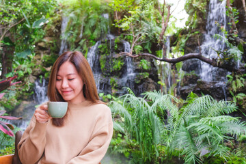 Wall Mural - Portrait image of a young woman holding and drinking coffee while sitting in the garden with waterfall