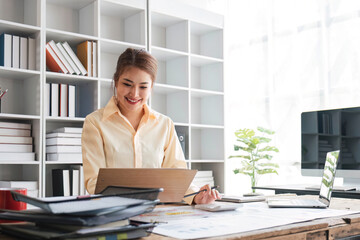 Wall Mural - Beautiful and happy young Asian businesswoman looking at her laptop screen while enjoying her morning coffee at her desk in the office.