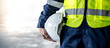 © Summer Paradive - Safety workwear concept. Male hand holding white safety helmet or hard hat. Construction worker man in protective suit and reflective green vest standing with building concrete floor in the background