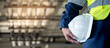 © Summer Paradive - Safety workwear concept. Male hand holding white safety helmet or hard hat. Construction worker man in protective suit and reflective green vest standing with building piping system in the background