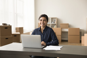 Canvas Print - Professional portrait of happy Indian entrepreneur girl sitting at workplace table in internet store warehouse, looking at camera, smiling, working at laptop computer