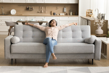 Cheerful positive Indian new homeowner stretching body on stylish comfortable couch in cozy home interior, smiling, laughing, enjoying leisure, relaxation, celebrating real estate buying