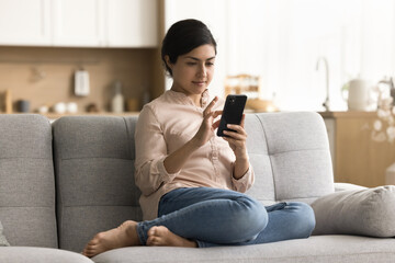 Relaxed focused young Indian woman sitting on cozy sofa, holding smartphone, typing message, using online application, Internet technology, enjoying leisure, entertainment on digital device