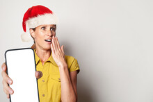 Excited Surprised Woman In Santa Hat Holding Smartphone With Empty Blank Display Screen And Present Box On White Background