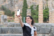 Woman smiling and taking a selfie photo at the sanctuary of Apollo at Delphi in Greece. A famous touristic destination.
