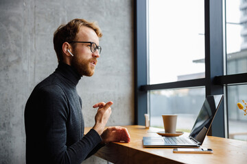 A young red-haired guy, a programmer or businessman in glasses, is sitting in a stylish cafe at a laptop and is talking through a webcam. Freelancer works remotely. Online communication.