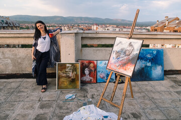 woman artist in the city, drinking coffee on a rooftop by her paintings and smiles