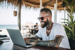 © Jasmina - Smiling handsome hipster man with tattoos sitting and working on his laptop in the beach bar