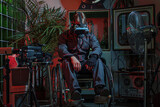 Fototapeta Tęcza - A man in overalls with a cyber helmet on his head, sitting in a wheelchair among old things, high technology and low standard of living of society, concept