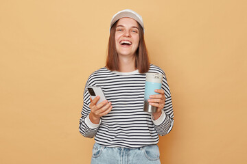 Wall Mural - Laughing woman wearing baseball cap and striped shirt holding thermos and using smart phone browsing internet enjoying isolated over beige background