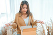 © oatawa - Bad delivery service, Dissatisfied young asian woman unpacking cardboard box while on armchair at home, Female feeling upset with wrong parcel