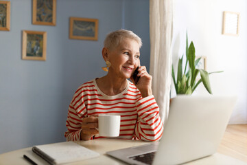 Cute stylish senior smiling female freelancer in striped shirt drinking tea and talking on phone during coffee break in online work at home office, sitting in front of laptop next to window