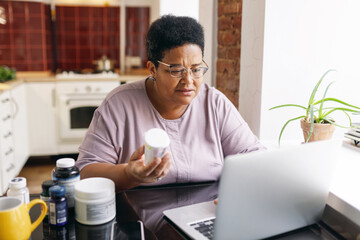 Horizontal image of black african american overweight elderly woman in spectacles holding bottle of food supplements and using laptop to order vitamins online sitting at kitchen table