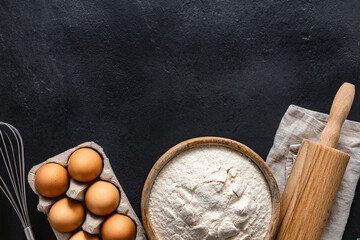 Poster - Flour in wooden bowl with eggs and rolling pin on a dark background top view. Organic ingredients for homemade baking