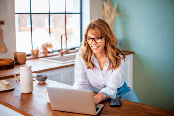 Wall Mural - A middle-aged woman standing at the kitchen counter in the morning and using a laptop