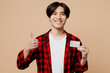Young smiling man of Asian ethnicity he wear red shirt casual clothes hold in hand mock up of credit bank card show thumb up isolated on plain pastel light beige background studio. Lifestyle concept.