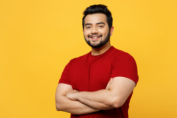 Side view young smiling happy fun Indian man he wearing red t-shirt casual clothes hold hands crossed folded look camera isolated on plain yellow orange background studio portrait. Lifestyle concept.