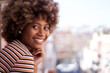 © CarlosBarquero - Young African American woman looking smiling on camera. Cheerful black afro girl leaning on home window. Portrait female beautiful expression posing for photo. Copy space and blurred background.