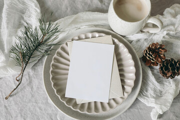 Wall Mural - Christmas still life. Blank greeting card, invitation mockup on ceramic plate. Pine cones and pine tree branches on white linen tablecloth. Cup of coffee, winter breakfast. Festive holiday flat lay