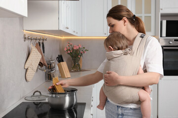 Wall Mural - Mother holding her child in sling (baby carrier) while cooking pasta in kitchen