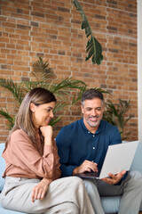 Smiling mature Indian business man manager having conversation with client or colleague at corporate meeting. Two happy professionals working using laptop talking sitting in office on sofa. Vertical