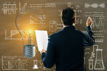 Back view of thoughtful young european businessman with paperwork and glowing light bulb on chalkboard wall background with mathematical formulas. Science, idea and education background.