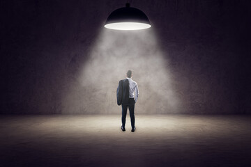 Back view of young businessman standing in spotlight of lamp in concrete interior.