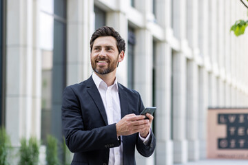 Young cheerful male businessman stands outside office buildings and uses the phone, reads news, types messages, waits for an appointment, looks to the side with a smile