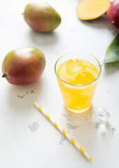 Wall Mural - Glass of mango juice with straw and fresh raw organic mangoes on light background.