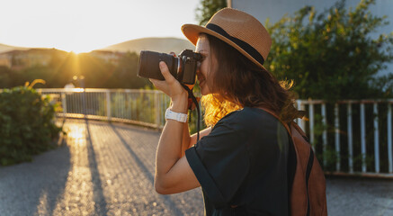 Wall Mural - Young caucasian woman traveler in a hat with a photo camera in her hands in a summer city at sunset