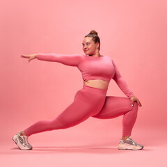 Full body portrait of young plus size sporty woman in pink top and legging training, stretching legs and hands isolated studio background.