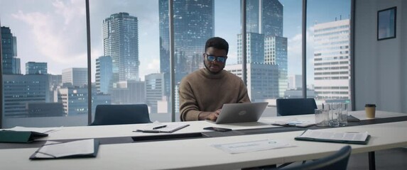 Poster - Confident Black Businessman Sitting at Desk in Modern Office, Using Laptop Computer, Next to Window with Big City with Skyscrapers View. Focused Sales Manager Planning Work Projects. Anamorphic Shot