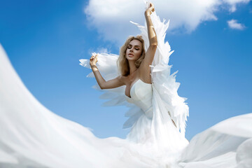 Beautiful sexy blonde woman in white long elegant dress and angel wings is sensually posing against the heaven on summer day. Angel woman.