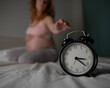 © Михаил Решетников - Caucasian pregnant woman sits on the bed and suffers from insomnia. Alarm clock in the foreground.