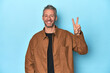 © Asier - Middle-aged caucasian man on blue backdrop joyful and carefree showing a peace symbol with fingers.