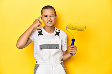 Painter woman, shaved head, with yellow roller in studio showing a disappointment gesture with forefinger.