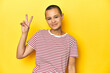 © Asier - Shaved head woman in red striped tee, yellow backdrop joyful and carefree showing a peace symbol with fingers.