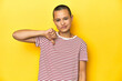 © Asier - Shaved head woman in red striped tee, yellow backdrop showing thumb down, disappointment concept.