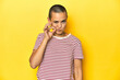 © Asier - Shaved head woman in red striped tee, yellow backdrop with fingers on lips keeping a secret.