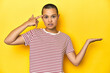 © Asier - Shaved head woman in red striped tee, yellow backdrop holding and showing a product on hand.