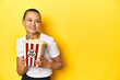 © Asier - Young woman eating popcorn, cinema concept, yellow backdrop