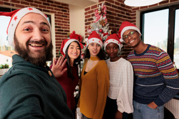 Wall Mural - Cheerful employees team wearing santa hats taking selfie while working in decorated company office. Coworkers posing for smartphone photo while celebrating christmas eve in coworking space