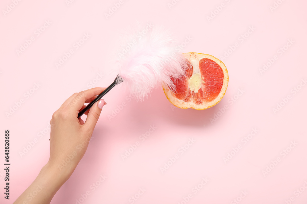 Female hand with feather stick and fresh grapefruit on pink background. Sex education concept