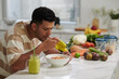 © DragonImages - Young hungry man eating muesli from bowl and having fresh juice or smoothie while sitting by kitchen table with fruits and vegetables