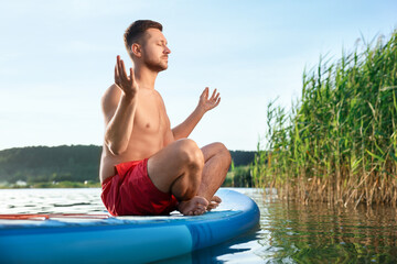 Wall Mural - Man meditating on light blue SUP board on river at sunset