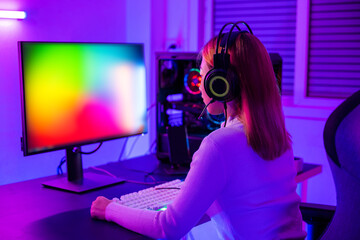 Wall Mural - Asian professional gamer playing online video game on desktop computer PC have colorful neon LED lights dark room, young woman in gaming headphones using computer for playing game at home, Back view