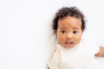 close-up portrait of a small African-American baby girl in a white bodysuit on a cotton bed at home, a six-month-old smiling joyful black baby